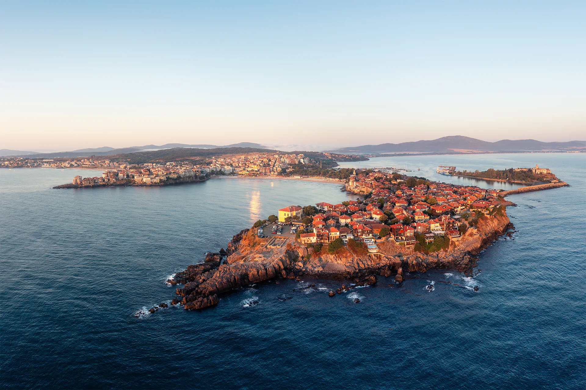 Aerial view of Sozopol Old Town on its rocky peninsula at sunrise, surrounded by the Black Sea, with the new town and harbour in the background.