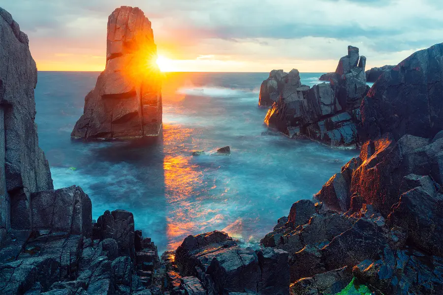 Sunrise at Palikari Rock near Sozopol, with the sun appearing beside the tall sea stack, long-exposure water and dark coastal rocks in the foreground.