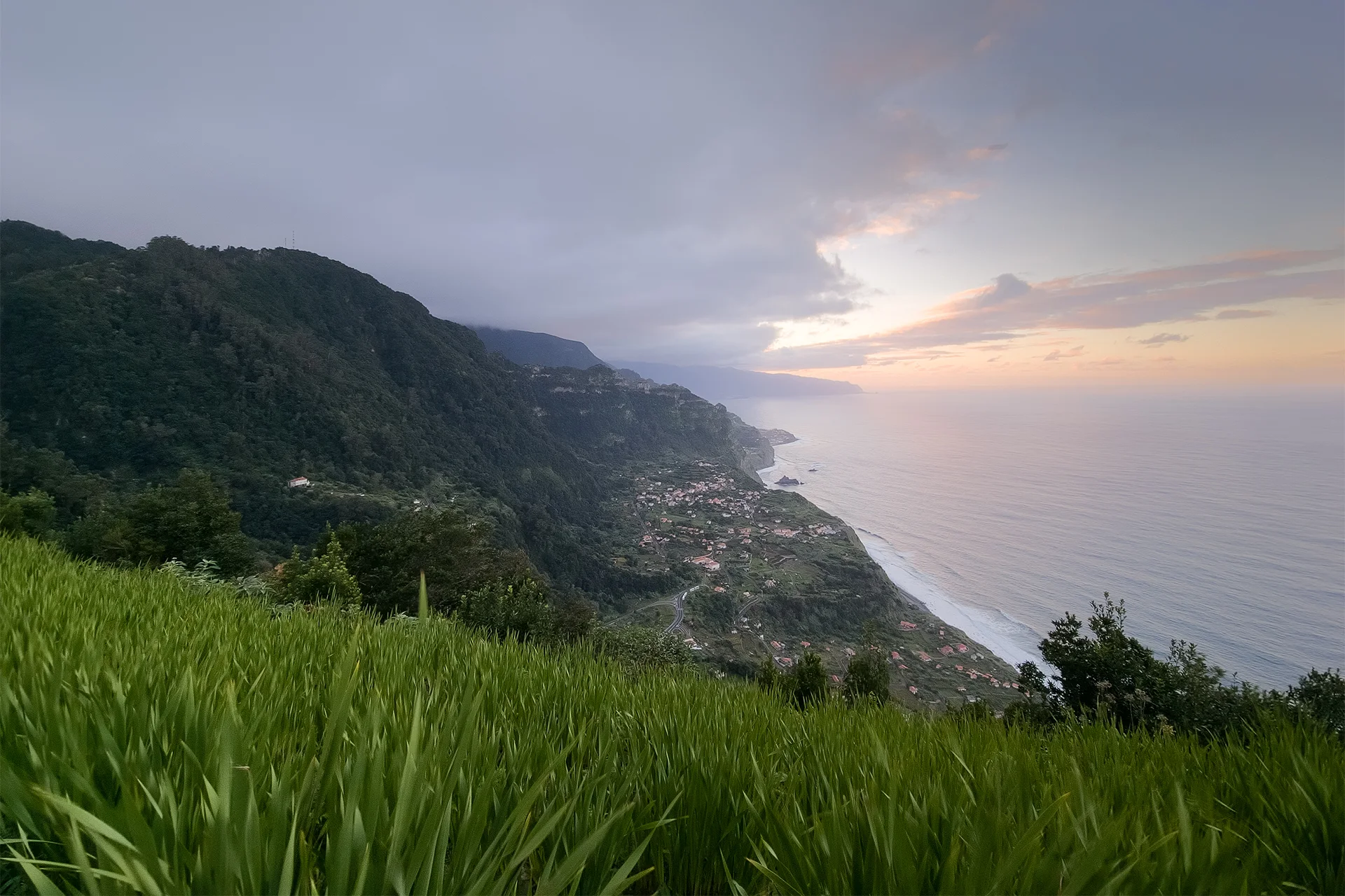 Sunset over Madeira’s northern coastline, with green mountain slopes, a coastal village and the Atlantic Ocean viewed from Miradouro da Ponta da Ladeira.