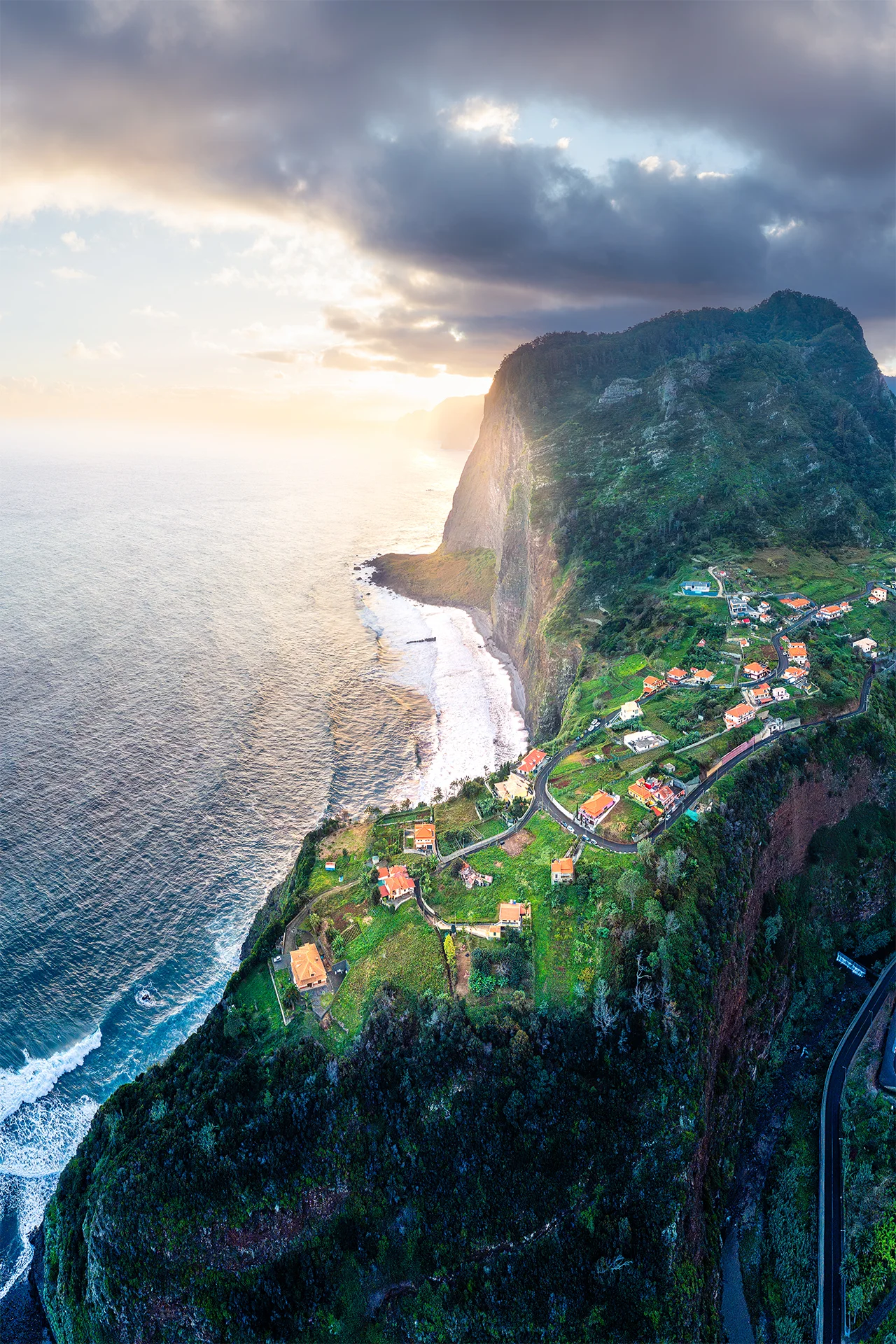 Aerial view of sunrise light over the Atlantic Ocean, steep green cliffs and houses near Miradouro do Guindaste, Madeira.