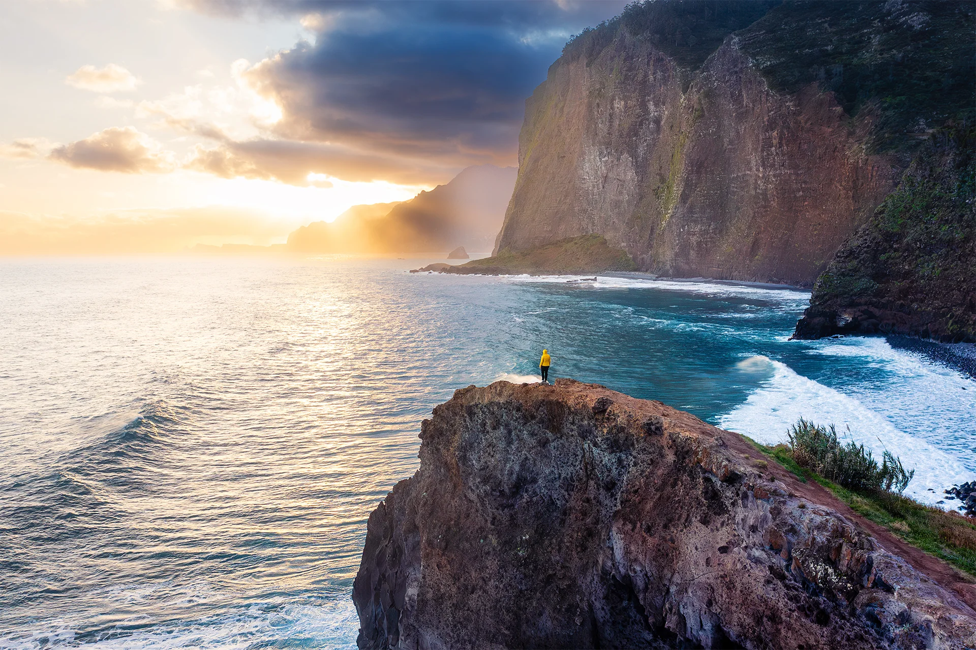 A person in a yellow jacket standing on a volcanic cliff above the Atlantic Ocean during sunrise at Miradouro do Guindaste, Madeira.