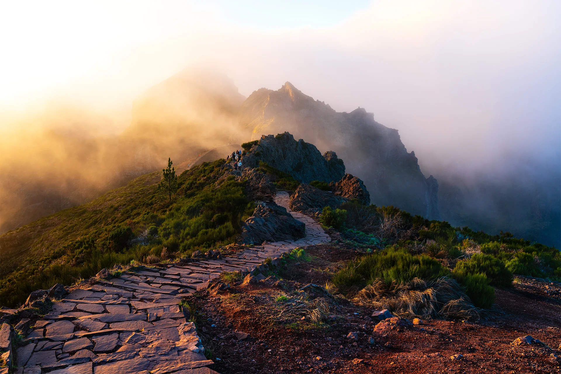 Sunlight breaks through moving clouds above the steep volcanic ridges and narrow mountain trail at Pico do Areeiro, Madeira.