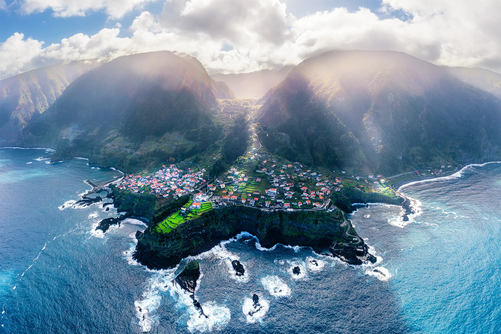 Aerial panorama of Seixal with red-roofed houses, cultivated terraces and steep green mountains rising above the Atlantic coast.