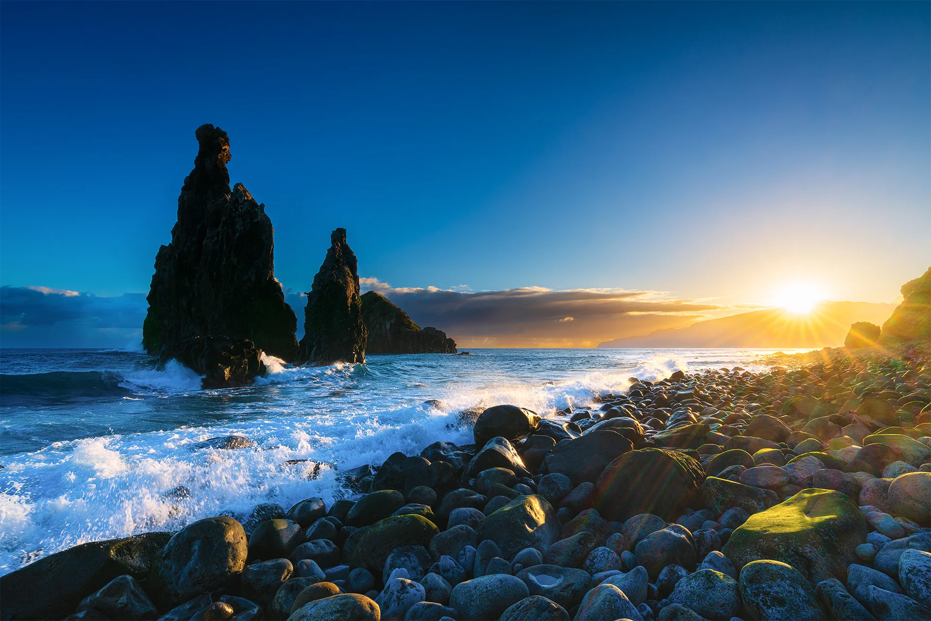 Sunrise over Ribeira da Janela, with Atlantic waves breaking around tall volcanic sea stacks and across the dark pebble beach.
