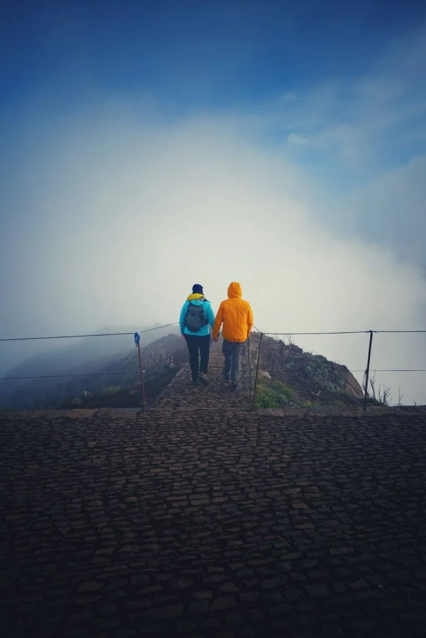 Two hikers in bright jackets walk along a stone mountain path into dense fog near Pico Ruivo, Madeira.