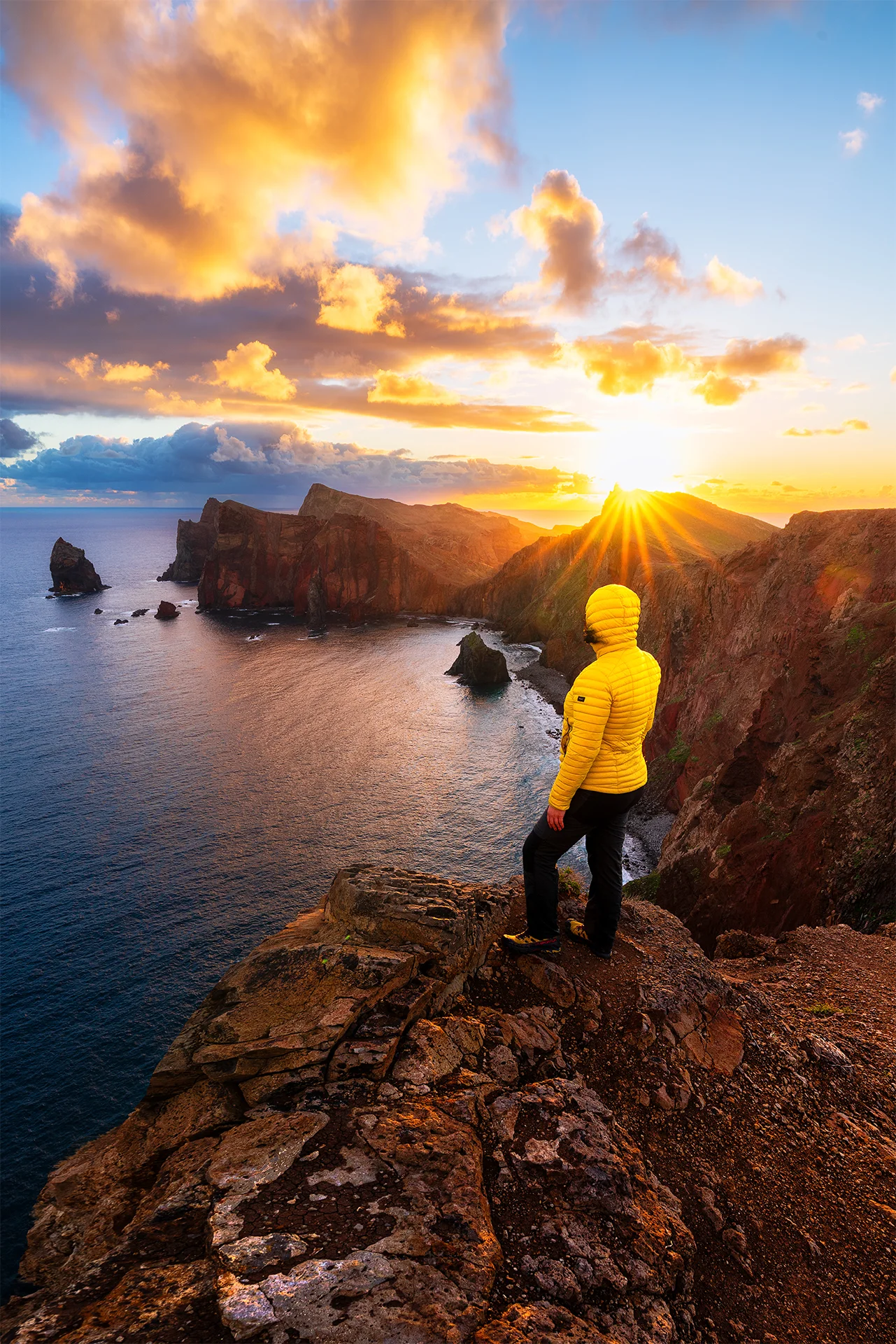 A person in a yellow jacket watches the sunrise over the rugged volcanic cliffs and Atlantic coastline at Miradouro da Ponta do Rosto, Madeira.