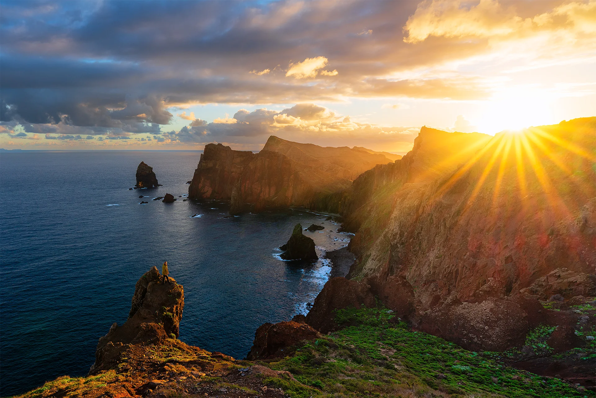 Golden sunrise over the rugged volcanic cliffs of Ponta do Rosto, with a lone person standing on a rocky outcrop above the Atlantic Ocean.