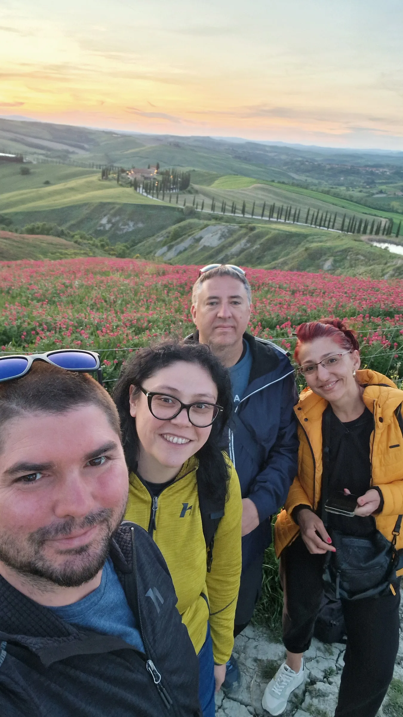 Four friends taking a group selfie at sunset above the rolling hills, cypress-lined roads and red flower fields of Tuscany.