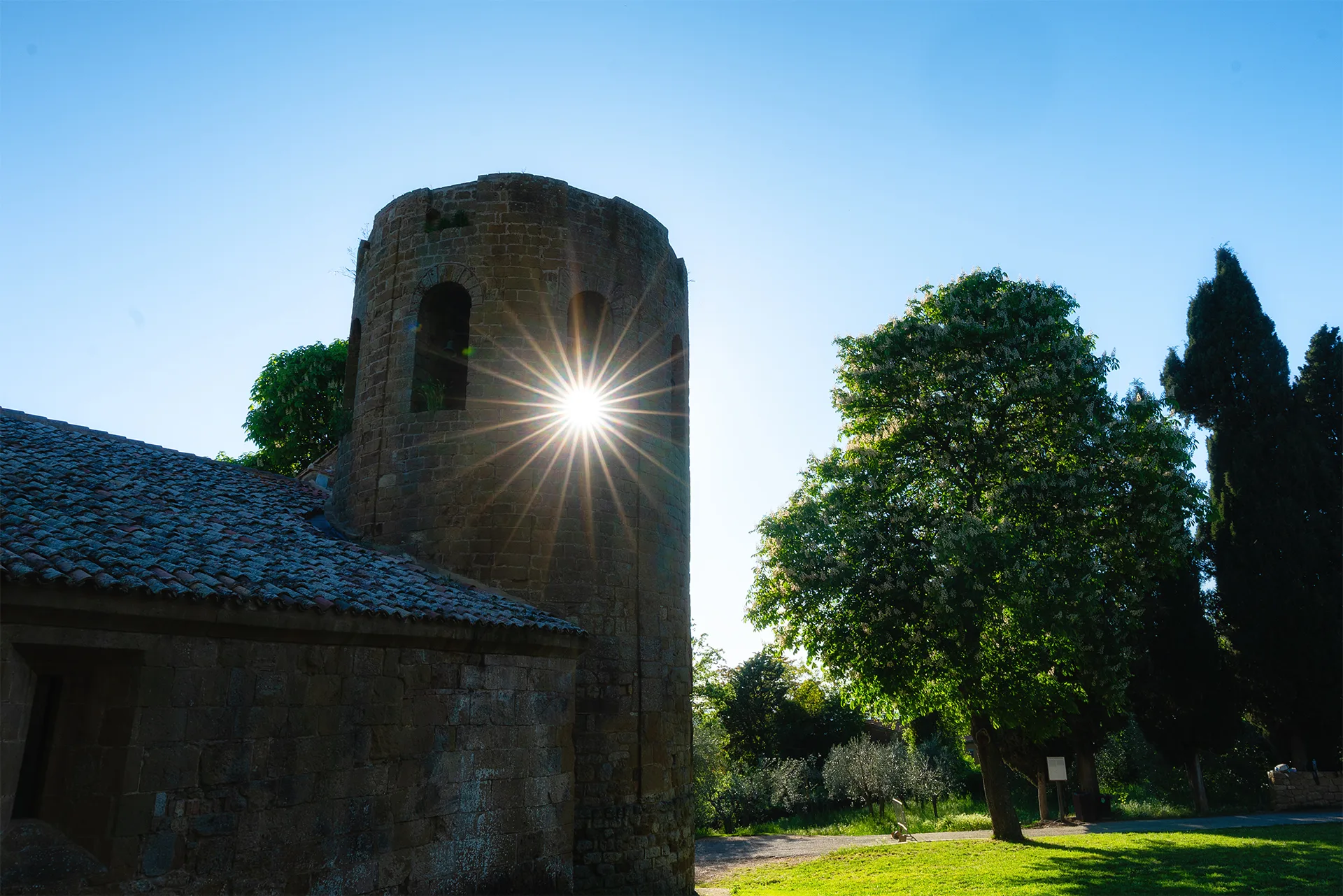 A bright sunburst shining through the stone bell tower of Pieve di Corsignano in Tuscany, surrounded by trees and grass.