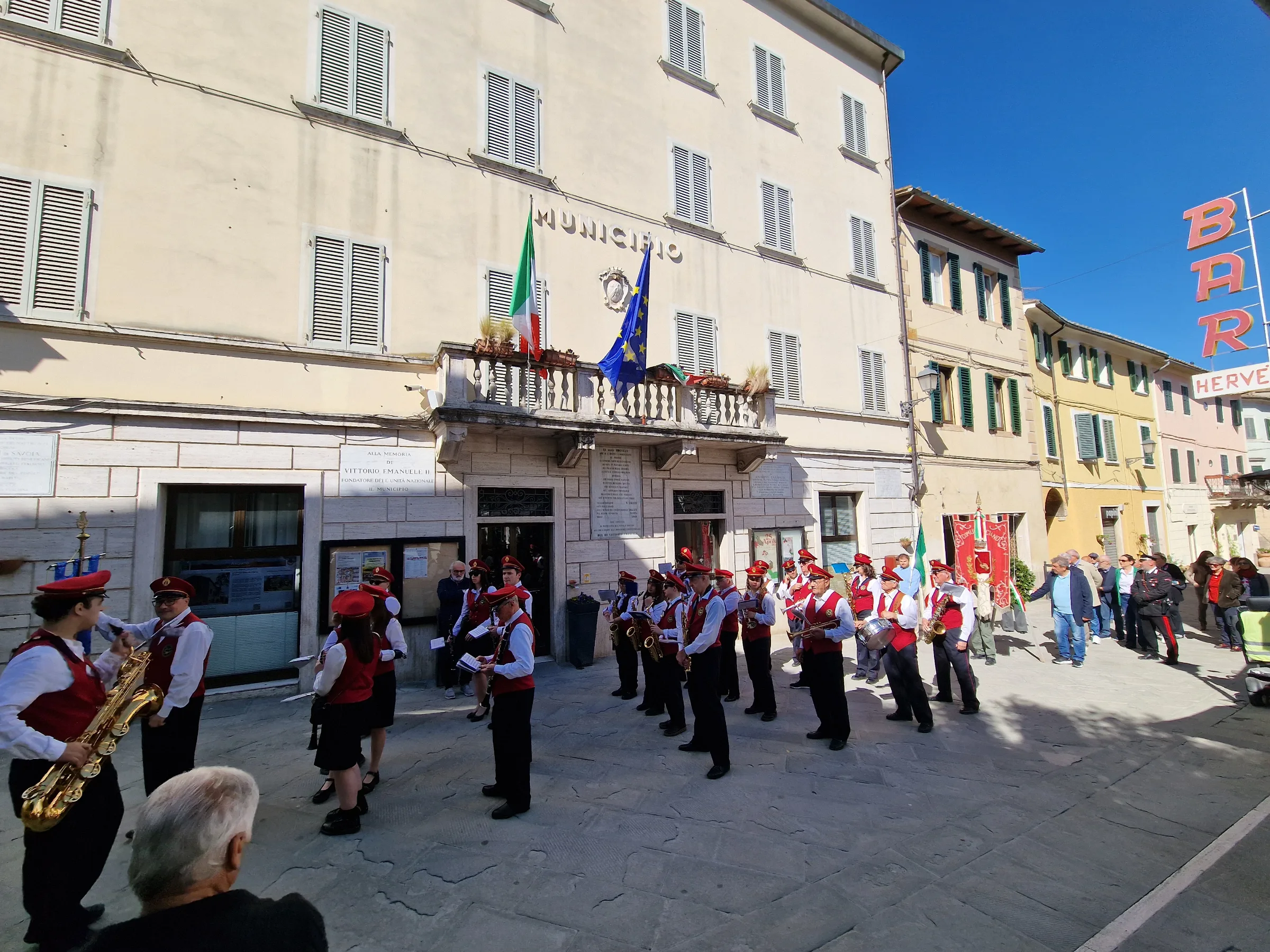 A brass band and local procession gathered in front of the town hall in Asciano, Tuscany, during a national holiday.
