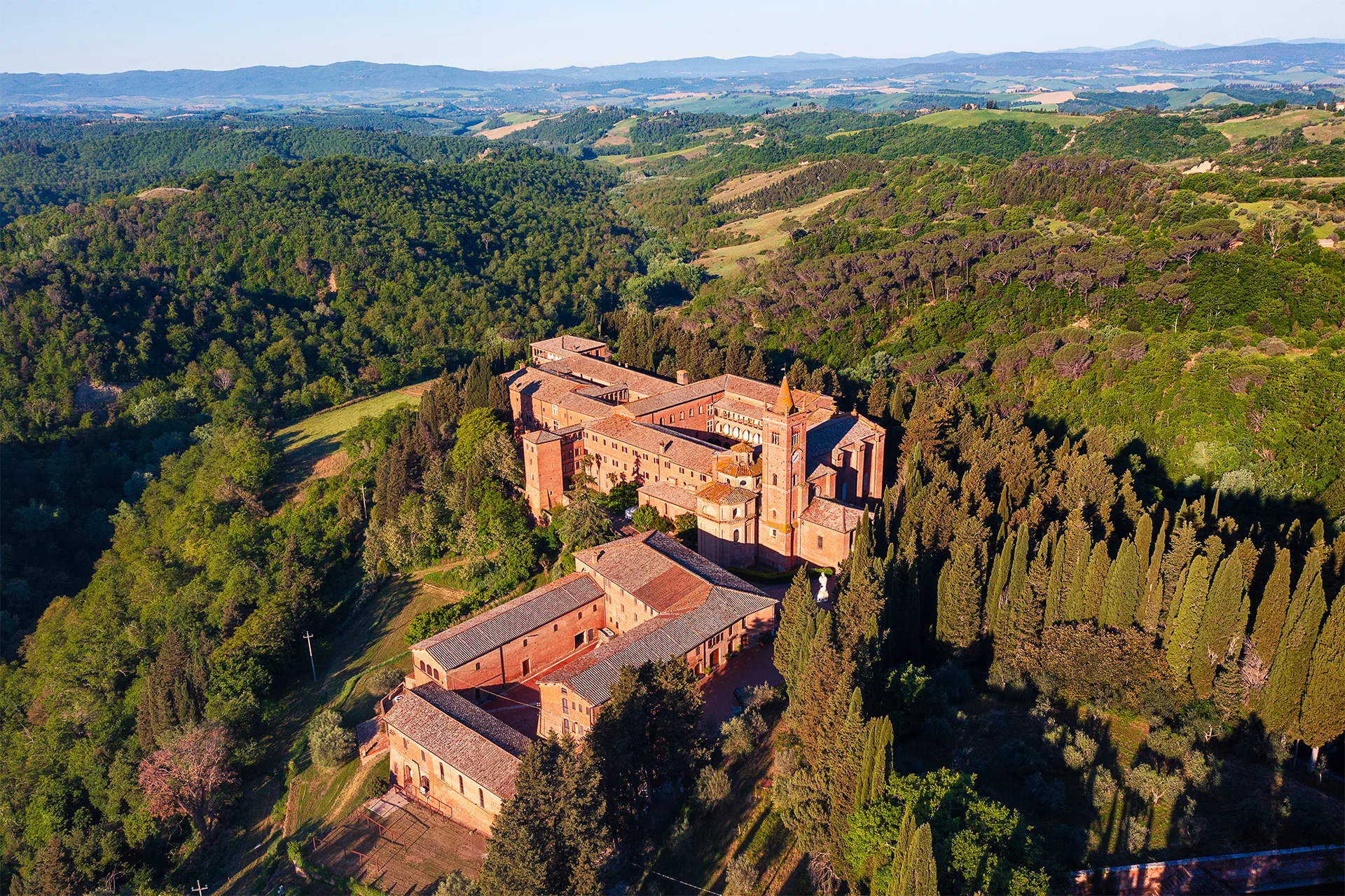 Aerial view of Abbazia di Monte Oliveto Maggiore surrounded by cypress trees, forests and rolling hills near Chiusure, Tuscany.