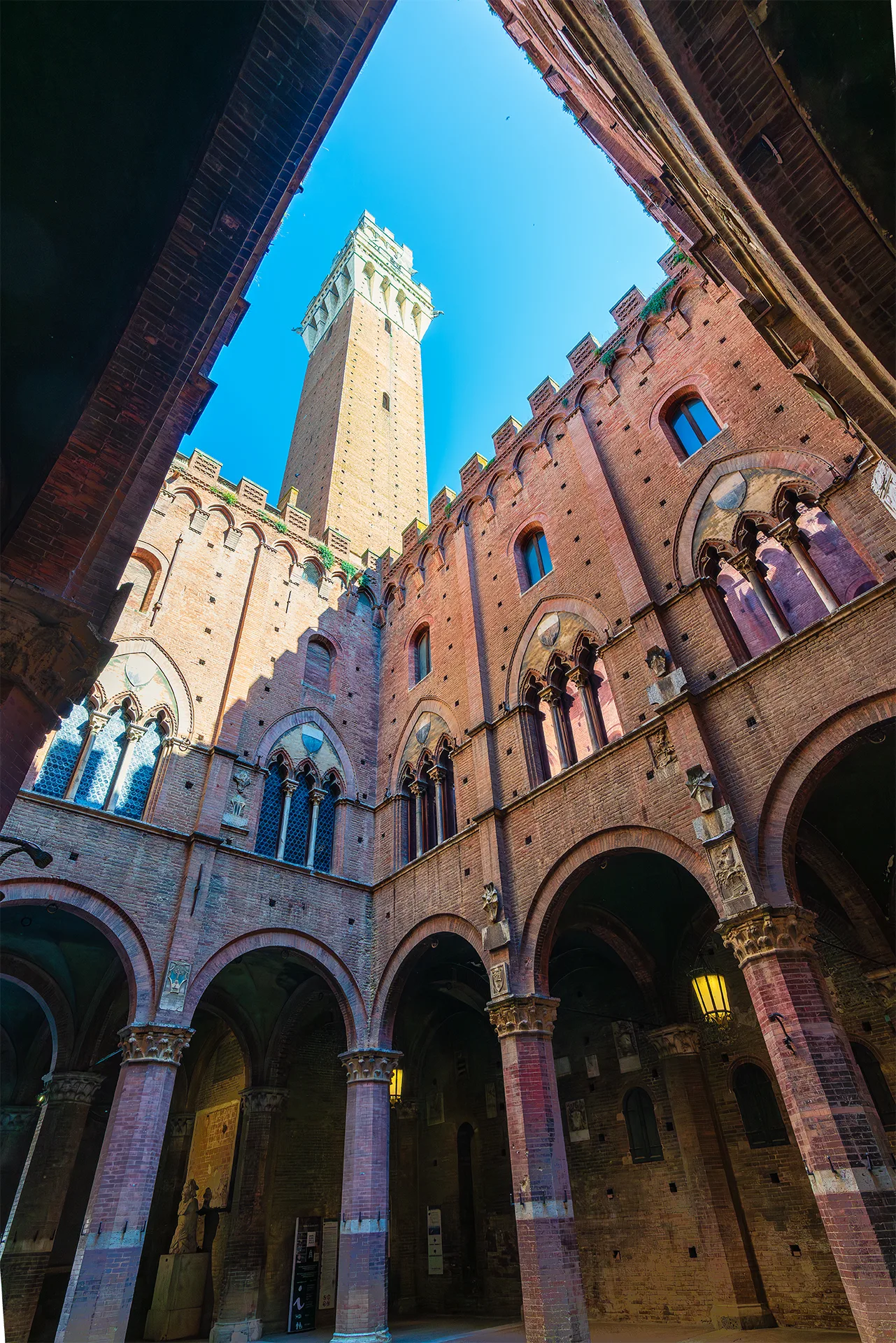 View from the inner courtyard of Palazzo Pubblico towards the tall brick tower and Gothic arches in Siena, Tuscany.