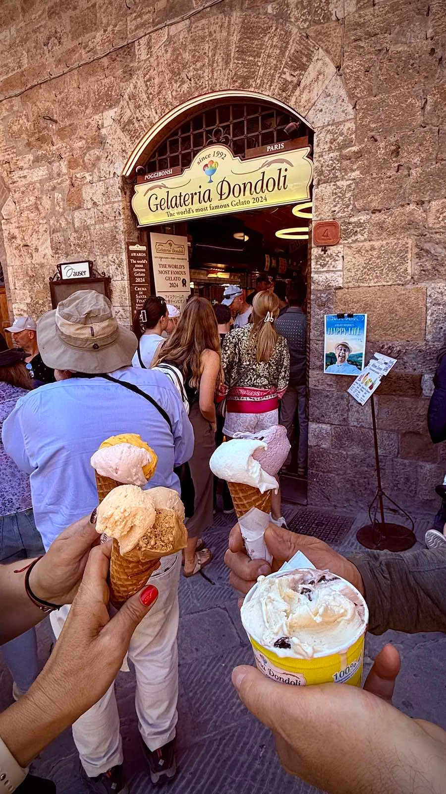 Friends holding gelato in front of the crowded Gelateria Dondoli in the historic centre of San Gimignano, Tuscany.