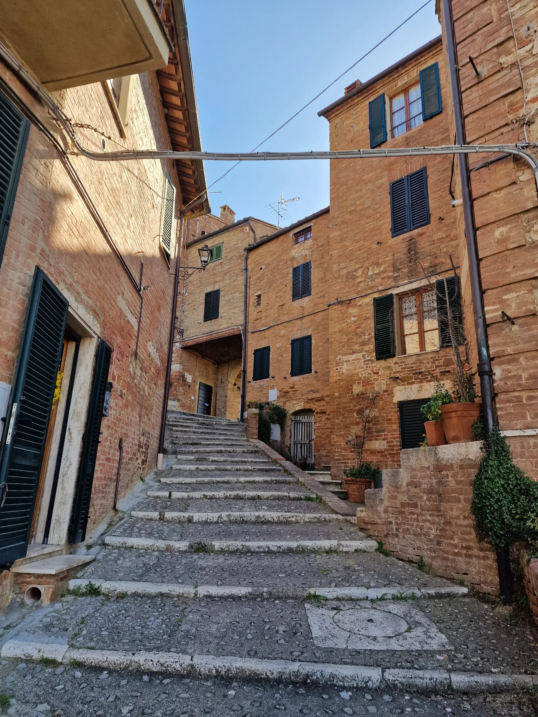 An old stone well surrounded by purple flowers and brick buildings along a quiet street in Chiusure, Tuscany.