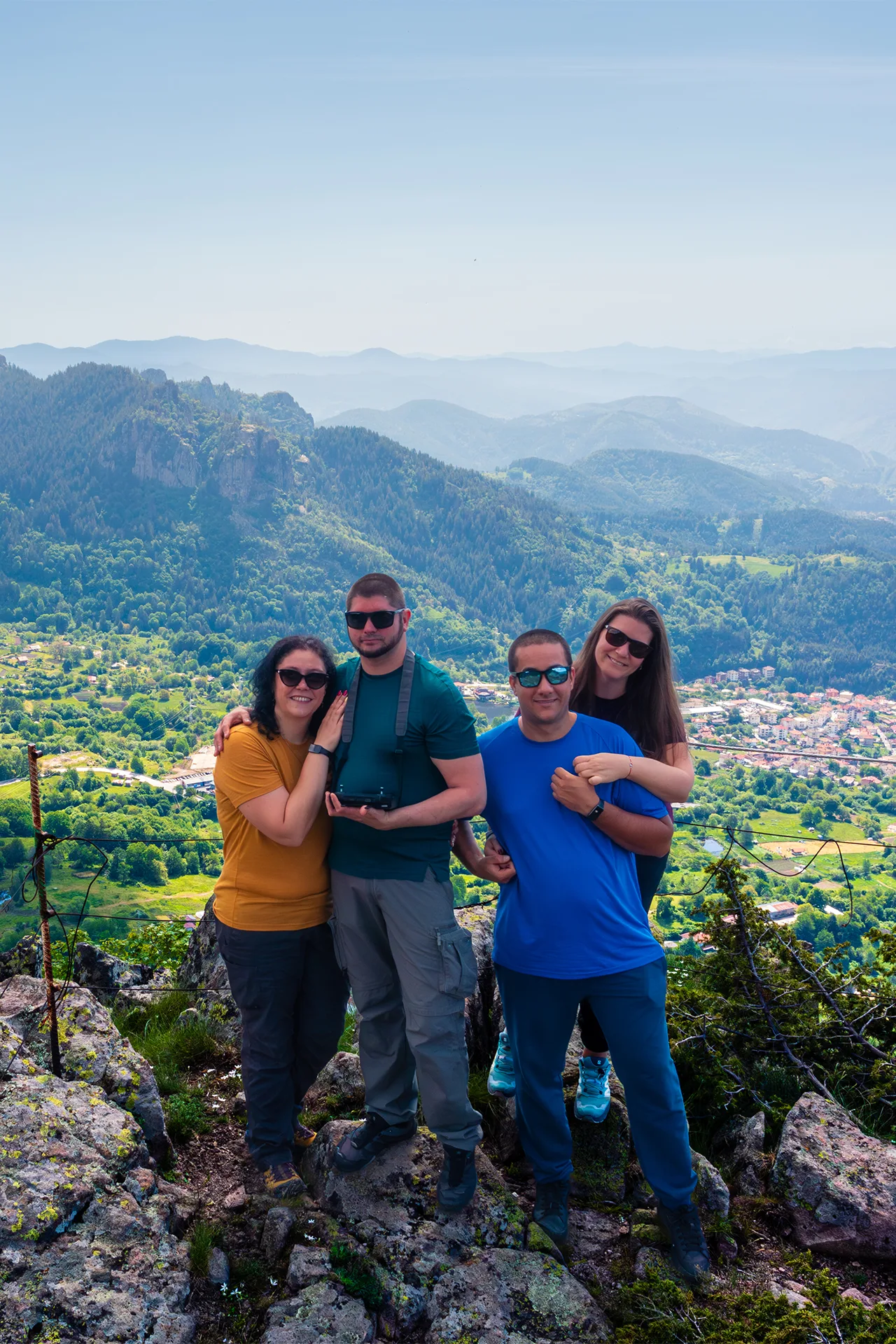 Four friends standing on the rocky viewpoint of Chervenata Skala above Smolyan and the forested Rhodope mountains.