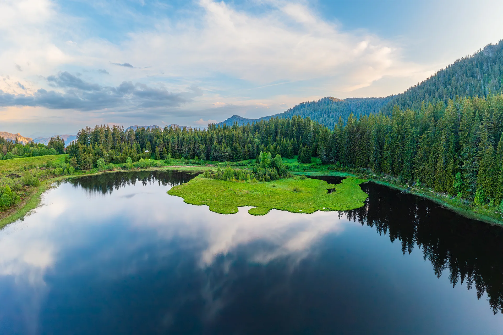 Aerial view of Lake Lagera reflecting the evening clouds, surrounded by green meadows and coniferous forest near Smolyan.
