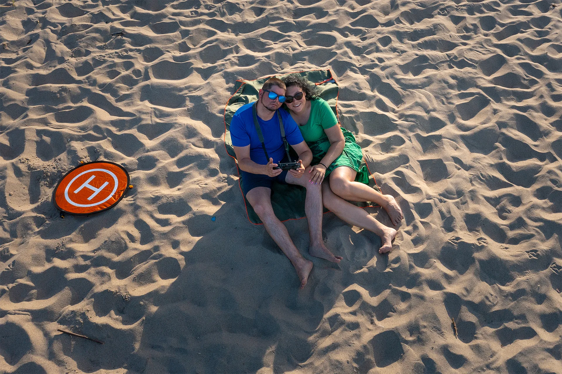 A couple sitting together on a blanket on a sandy beach, photographed from above while holding a drone controller.