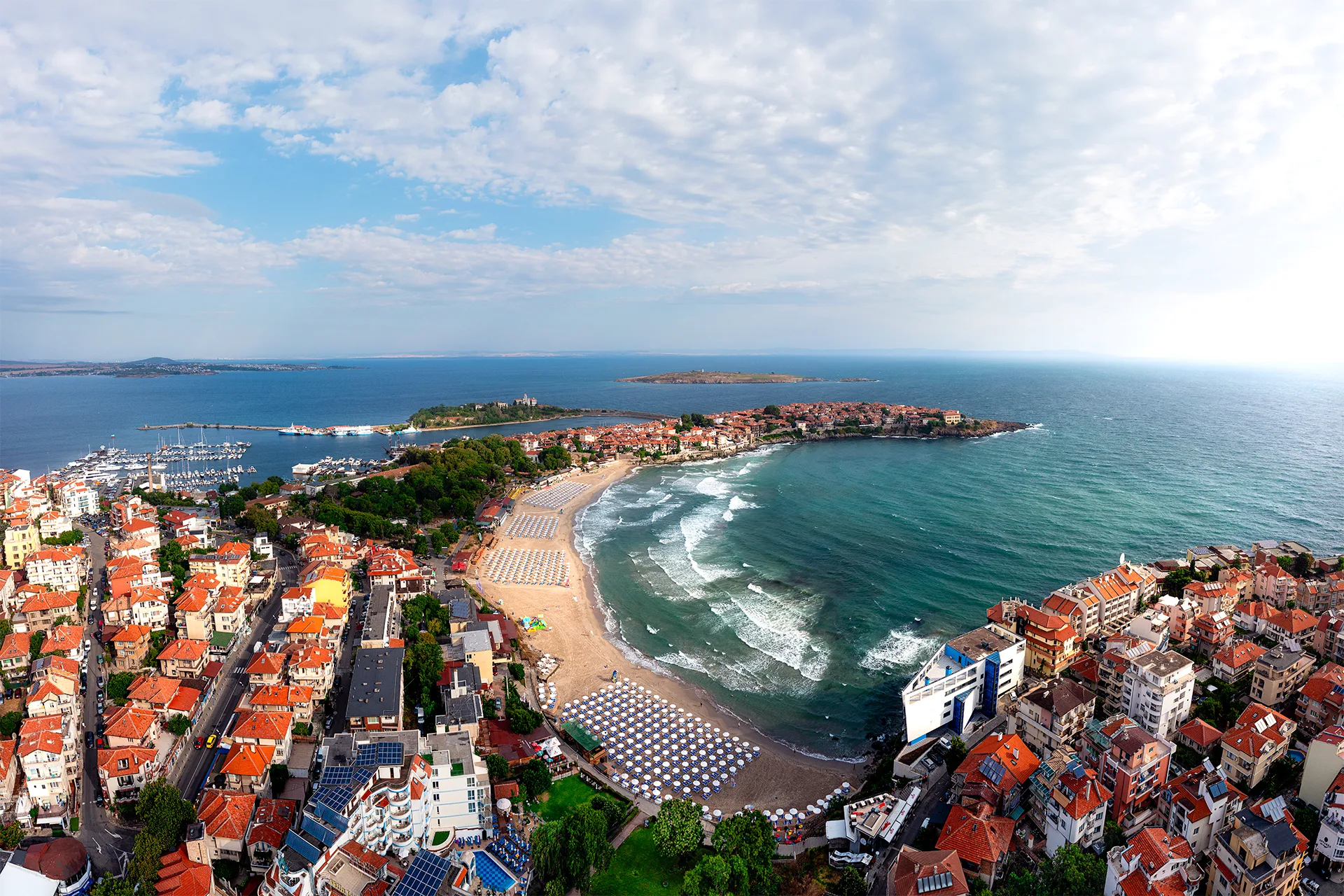 Aerial panorama of Sozopol, with the town beach, the Old Town, the marina and the Black Sea beneath a wide cloudy sky.