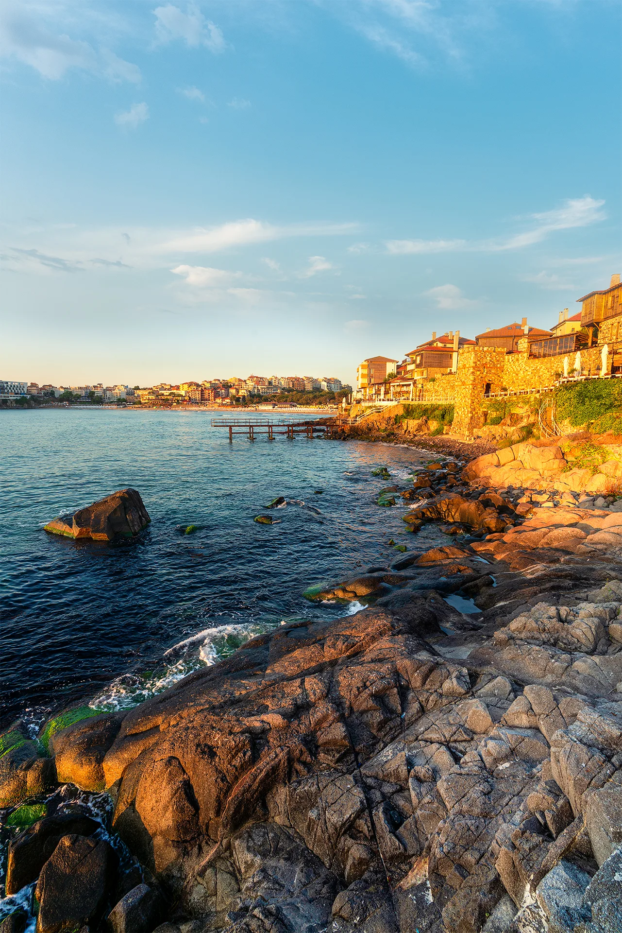Rocky shoreline below the old houses and stone walls of Sozopol, lit by warm morning sunlight beside the calm sea.