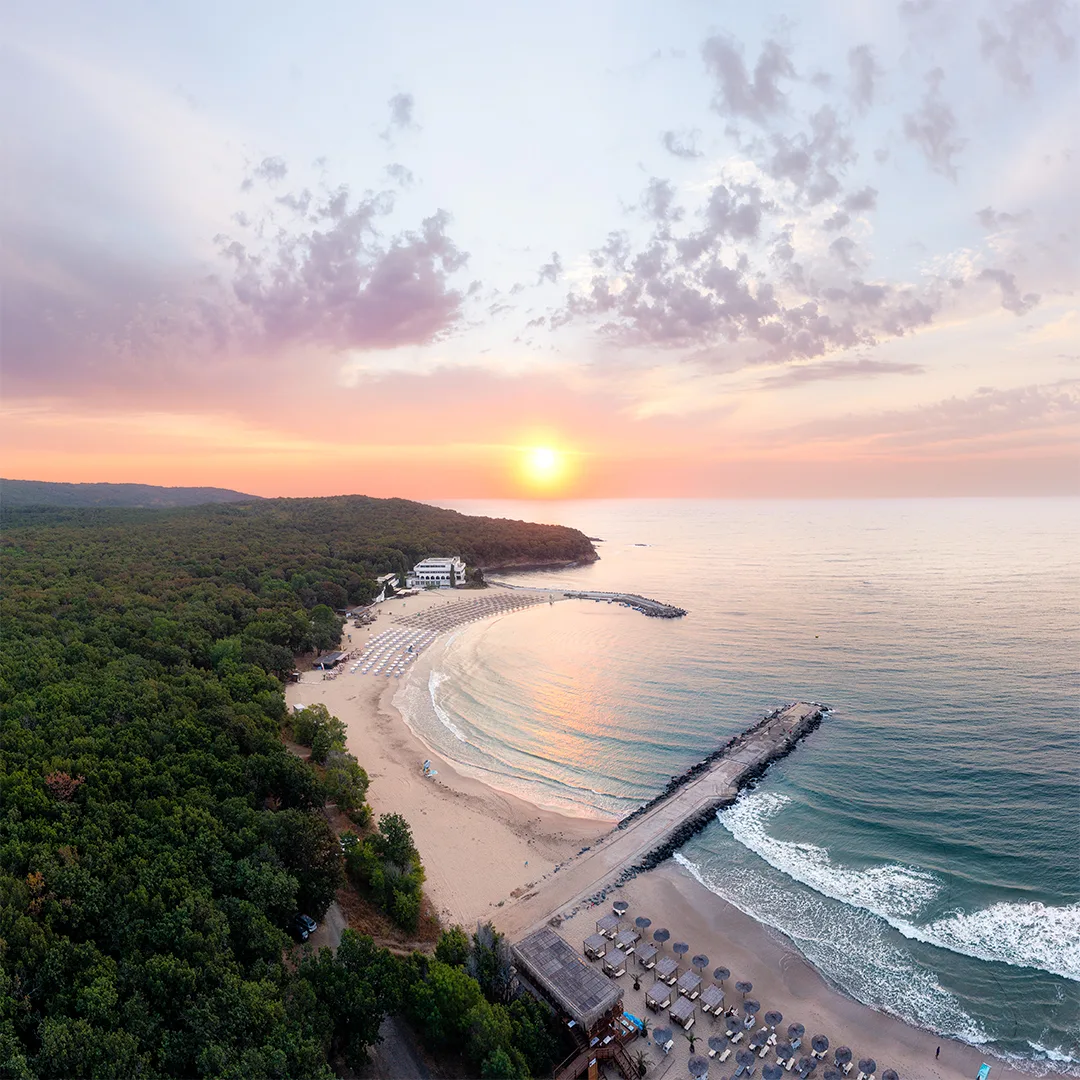 Aerial view of a sheltered sandy bay at sunrise, surrounded by dense forest, with calm water and two breakwaters extending into the sea.
