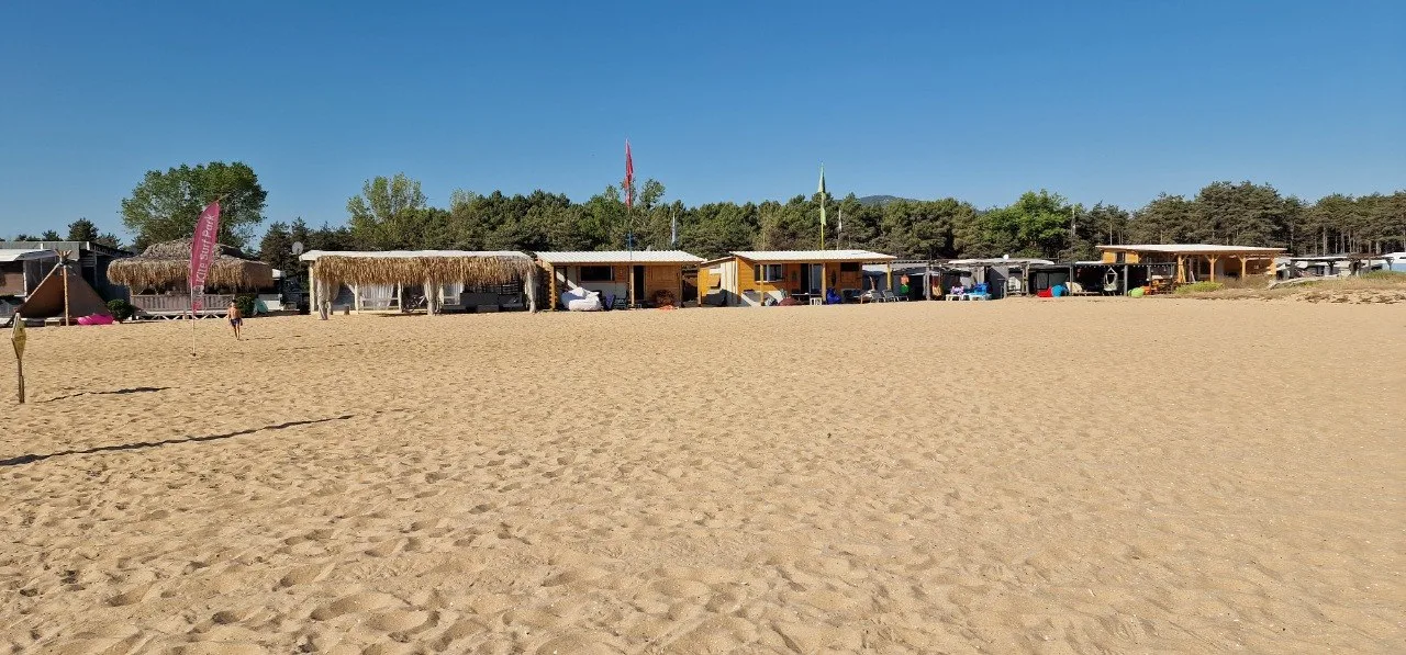 Wooden beach bungalows and shaded structures lined along a wide sandy beach, with trees behind them under a clear blue sky.