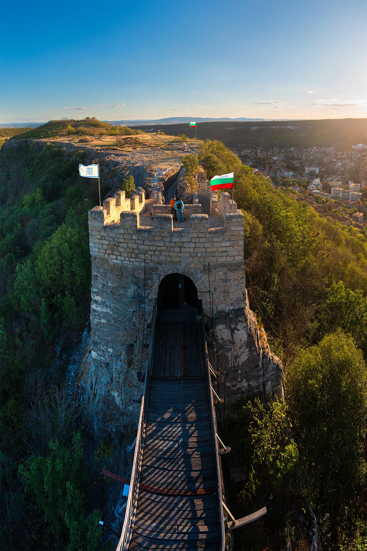 Stone tower and wooden bridge of Ovech Fortress above Provadia, with Bulgarian flags and the town visible in the valley below.