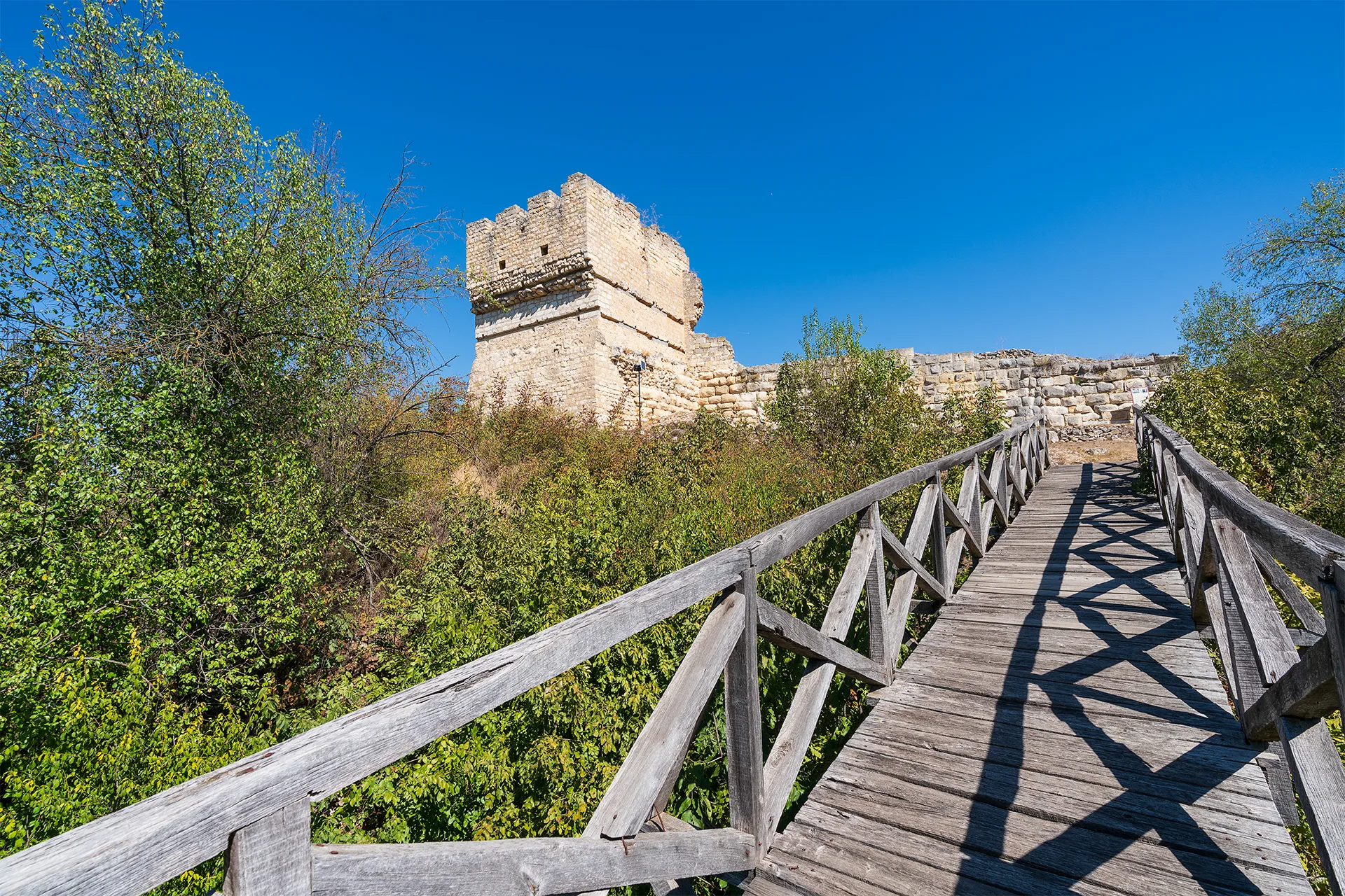 A wooden footbridge leading toward the preserved medieval stone tower of Cherven Fortress, surrounded by dense greenery under a clear blue sky.