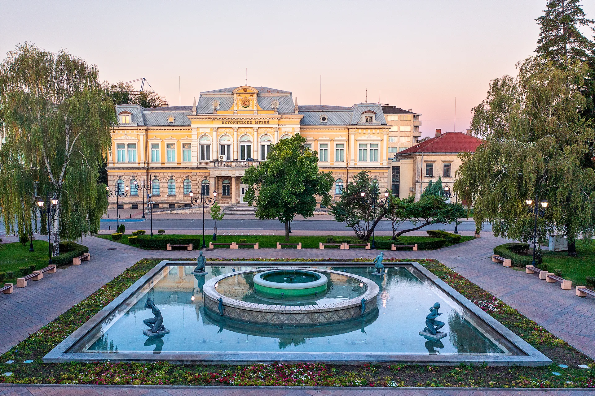 Regional Historical Museum in Ruse seen across a landscaped park with a fountain, sculptures, trees and flower beds.