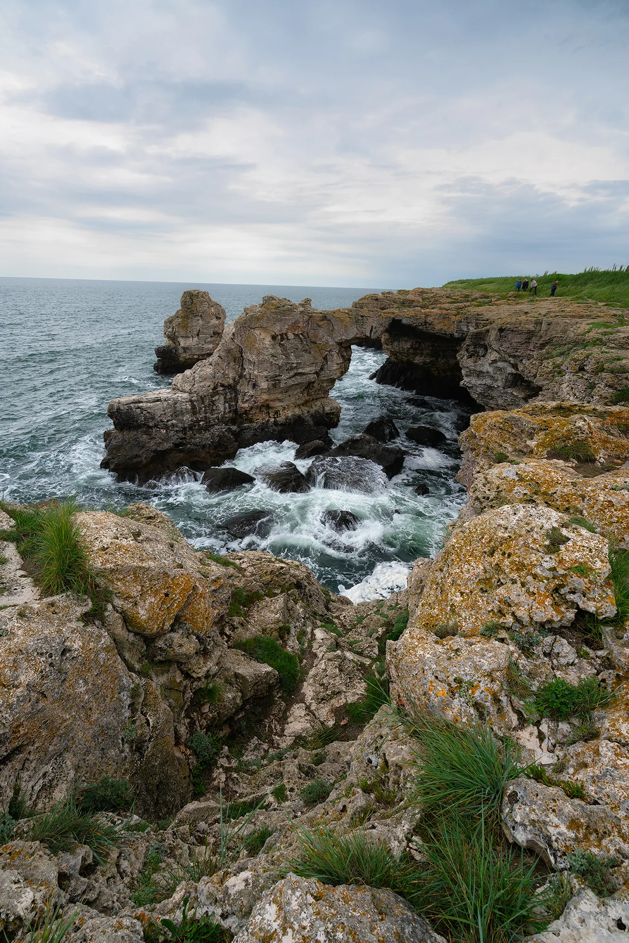 Rock arch on the coast of Tyulenovo with waves breaking below the cliffs under a cloudy morning sky.