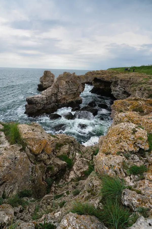 Rock arch on the coast of Tyulenovo with waves breaking below the cliffs under a cloudy morning sky.
