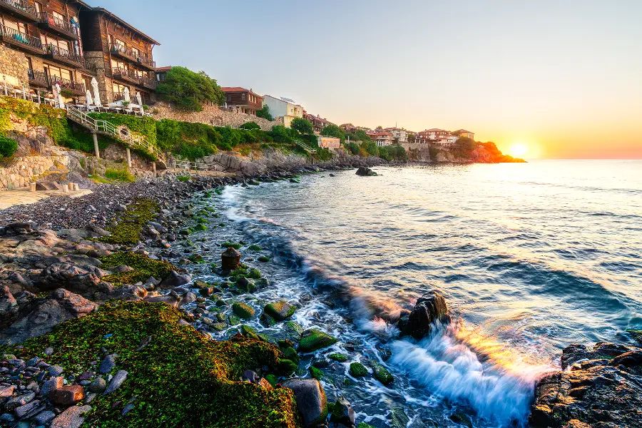 Sunrise over the Black Sea beside the rocky shore and traditional houses of Sozopol Old Town, Bulgaria.