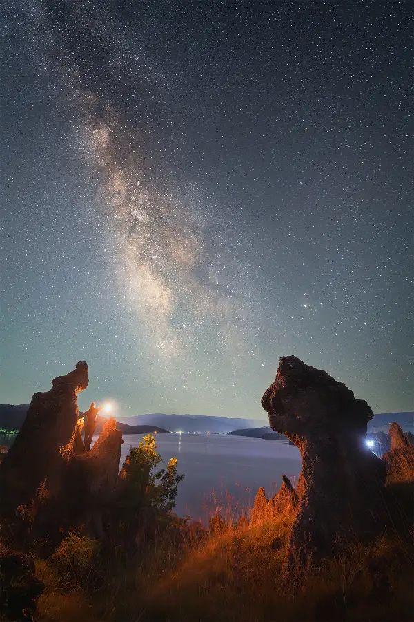Milky Way above rock formations on the shore of Pchelina Reservoir, with a person holding a light between the rocks and the water in the background.